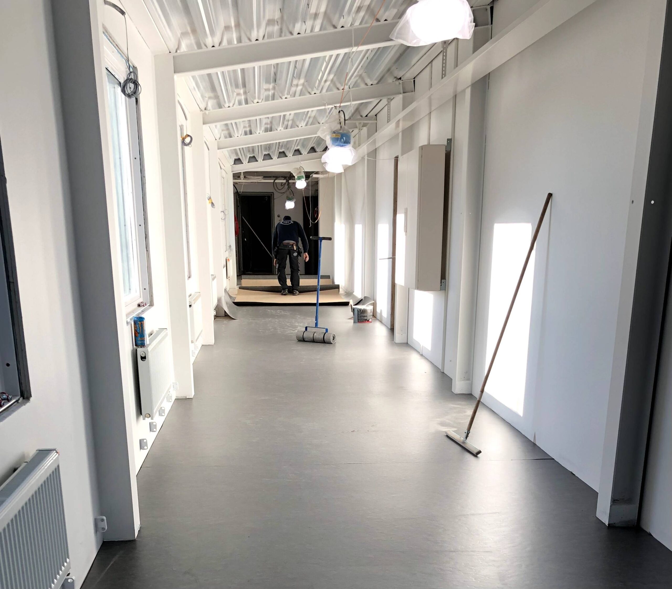 En lång, smal korridor med vita väggar och ett mörkt golv. En person arbetar i bakgrunden med en kraftig valsrullare. En sopborste står lutad mot väggen på höger sida. Det finns belysning och föremål utspridda längs väggarna.