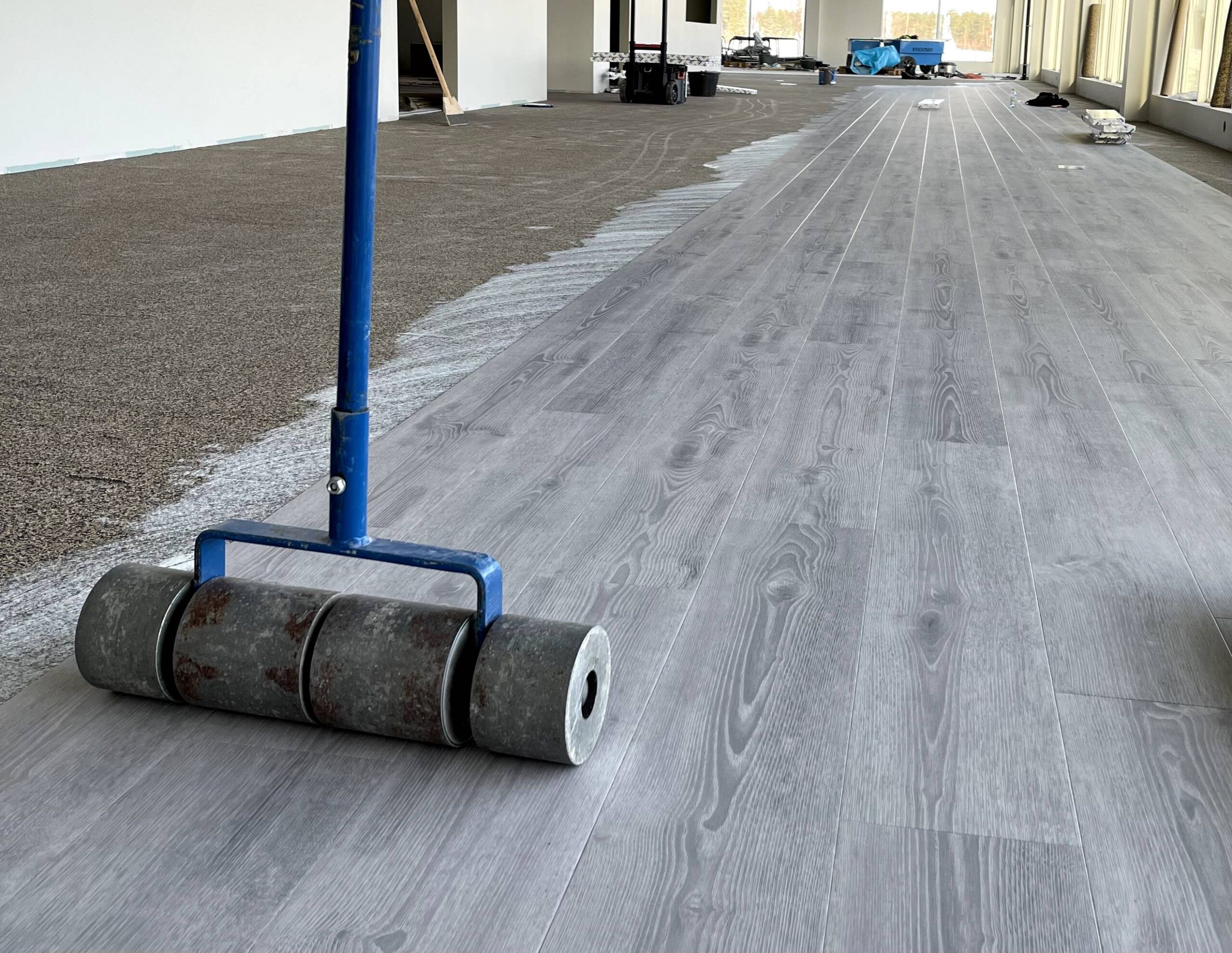 En stor rulleverktyg används för att lägga ett grått trägolv i ett stort rum, där halva golvet redan är installerat. Möbelplast och annan utrustning syns i bakgrunden.