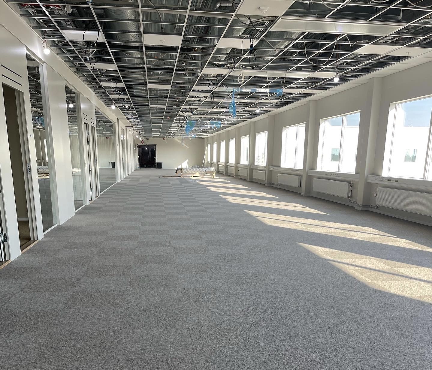 Ett stort, ljust rum under renovering, med exponerade ventilationsrör i taket och grått heltäckningsmatta på golvet. Flera fönster längs en vägg släpper in dagsljus.