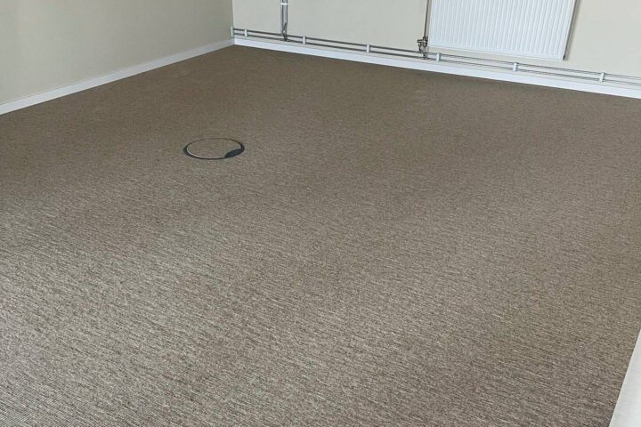 En tomt rum med en beige heltäckningsmatta, ett runt golvuttag i mitten och en radiator på väggen.