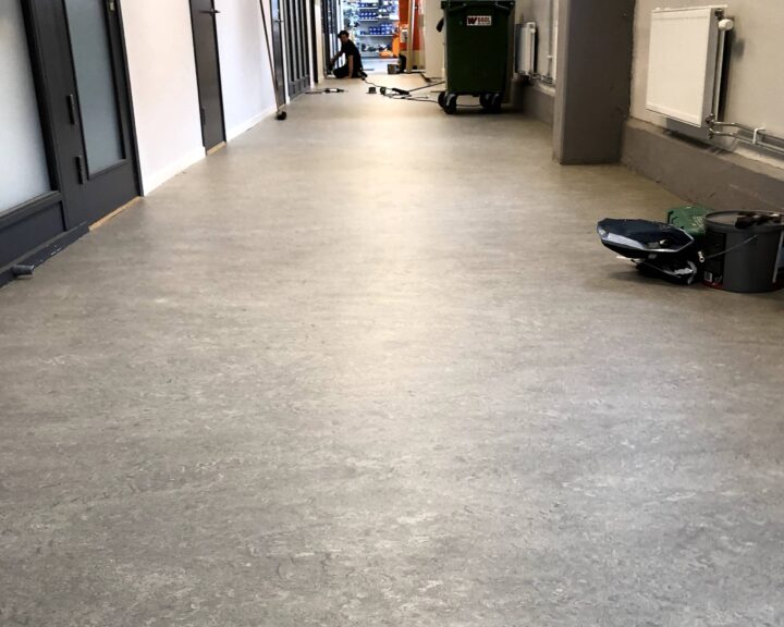 En lång, ljus korridor med stängda dörrar på ena sidan och radiatorer på den andra. I fjärran syns en person och en grön soptunna. Det finns målartillbehör på golvet.