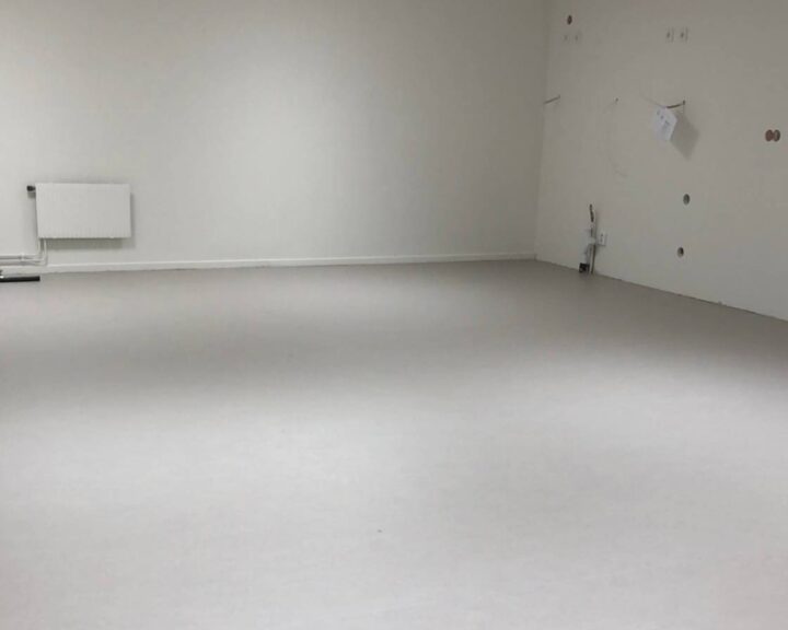 Ett tomt, ljust rum med ljusgrått golv, vita väggar och ett element på ena väggen. Ventilationsöppningar och eluttag syns.