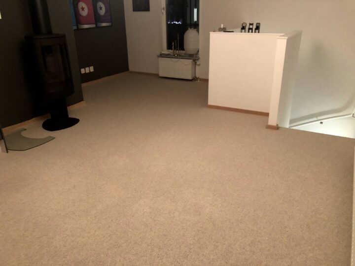 Ett rum med beige heltäckningsmatta, en svart kamin i hörnet och en vit vägg på höger sida. Flera föremål står ovanpå väggen.