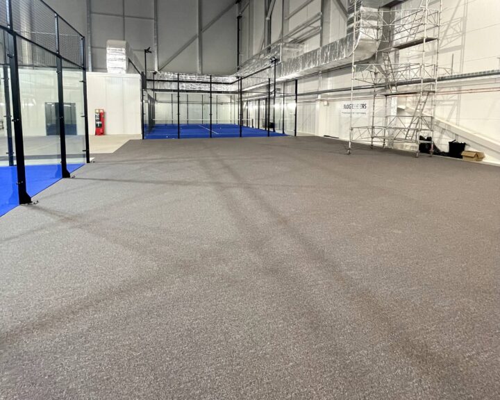 Inomhus sportanläggning under konstruktion med grått golv och blå banor. En byggnadsställning står till höger.