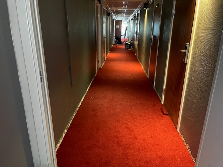 En lång korridor med röd heltäckningsmatta och dörrar på båda sidor. En person syns vid änden av korridoren.