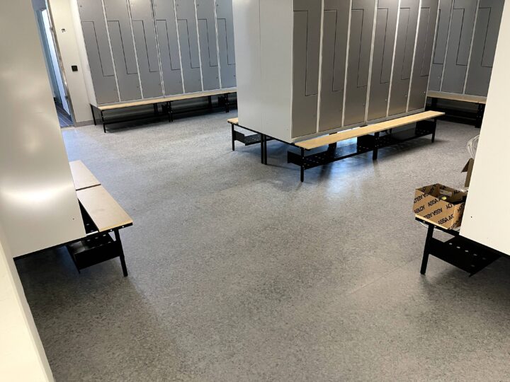 Bild av ett omklädningsrum med grå skåp och bänkar monterade på svarta metallben. Det finns en kartong på golvet.